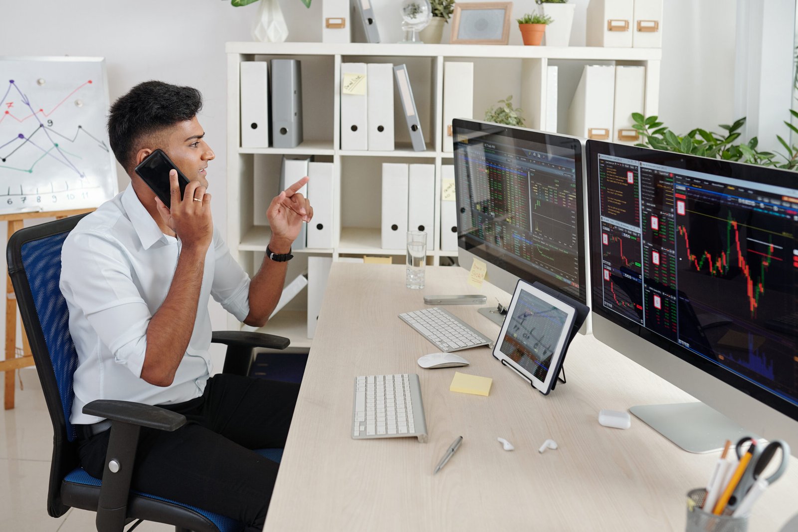 Positive Indian trader analyzing stock data on computer screens and consulting clients on phone